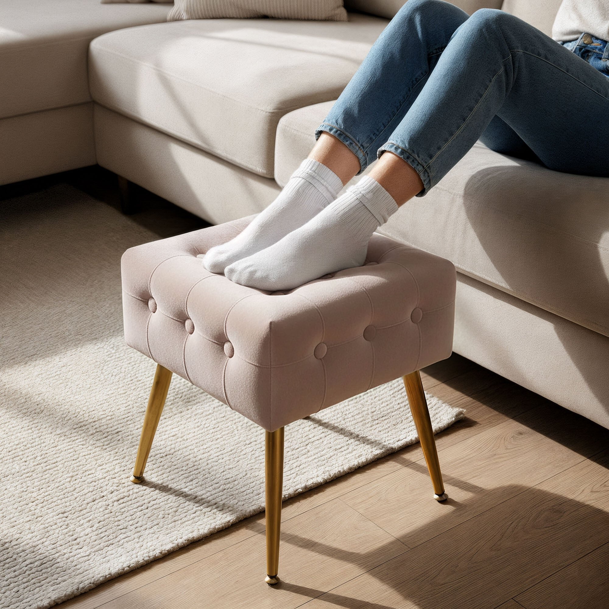 Pufa tapicerowana welurowa w kolorze pudrowego różu z pikowanym wzorem i złotymi nogami. Na pufie spoczywają stopy w białych skarpetkach, a w tle widoczna jest jasna sofa.