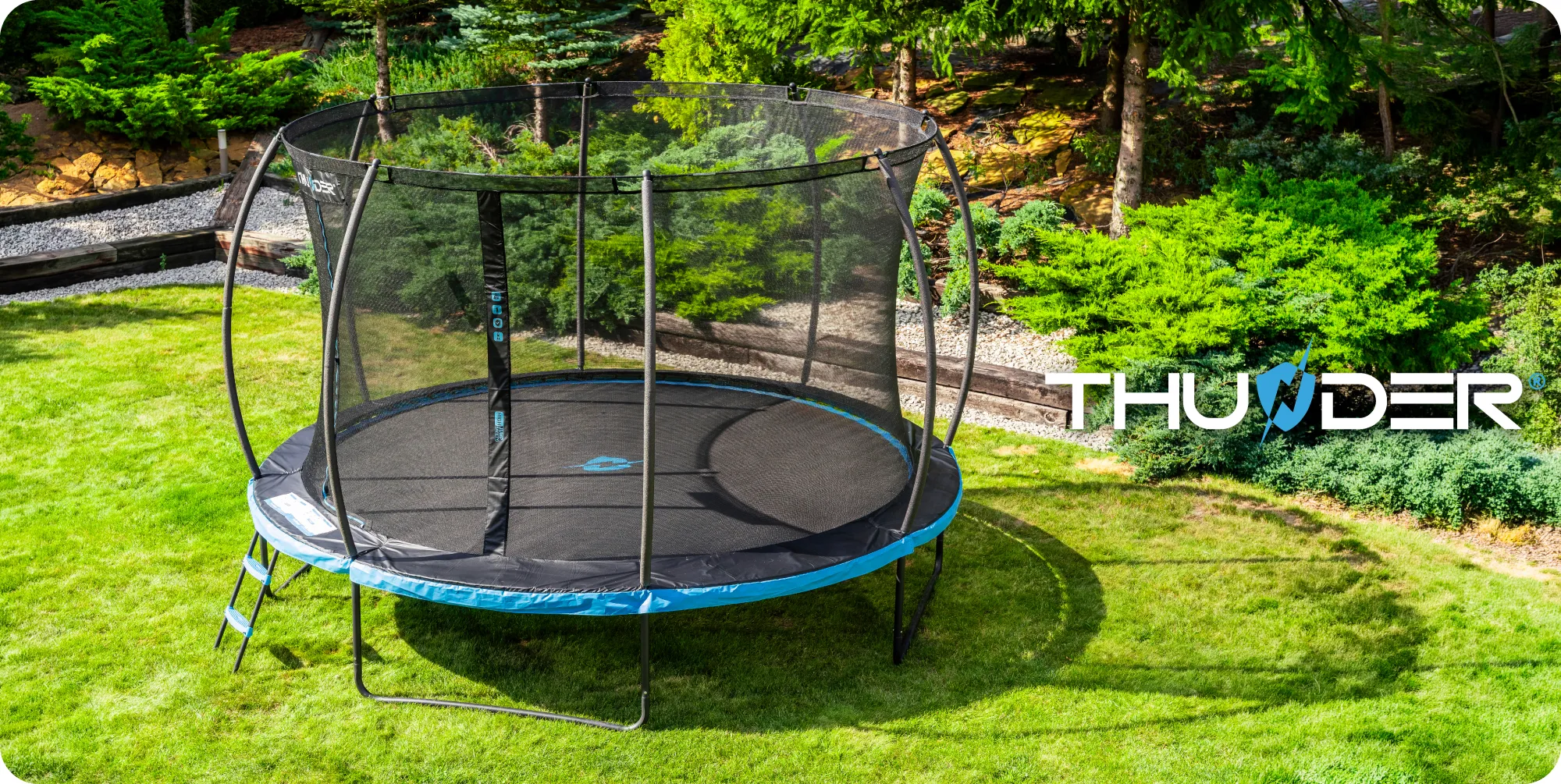 Na obrazie widoczna jest trampolina ogrodowa z siatką ochronną i drabinką, ustawiona na trawniku. W tle znajdują się drzewa i krzewy, a na trampolinie widnieje logo \'THUNDER\'.