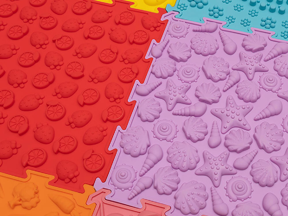 Mata sensoryczna ORTOPEDEK 128948 24.5 x 24.5 cm (12 elementów) Wielokolorowy Detal pastelowego, różowego elementu maty ortopedycznej Ortopedek. Zbliżenie pokazuje wypukłe wzory w kształcie stóp z logotypem marki, zaprojektowane do stymulacji receptorów dotykowych.