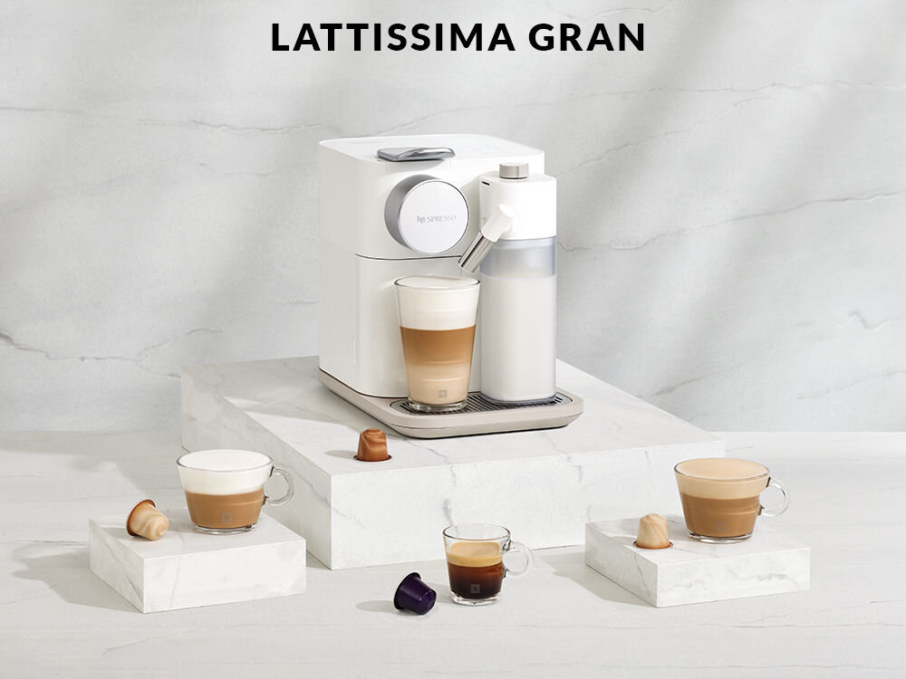 Na obrazie znajduje się biały ekspres do kawy z logo \'Nespresso\', obok którego stoją filiżanki z kawą i kapsułki. W tle widoczny jest napis \'LATTISSIMA GRAN\'.