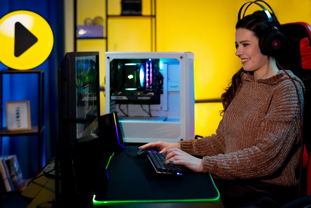 Komputer MAD DOG GeForce RTX5070 DLSS 4 Reflex Edition 2 ENDORFY500ARGB-WH-01WR32WP Uśmiechnięta kobieta w słuchawkach siedząca przy biurku i korzystająca z klawiatury przed dwoma monitorami oraz białym komputerem