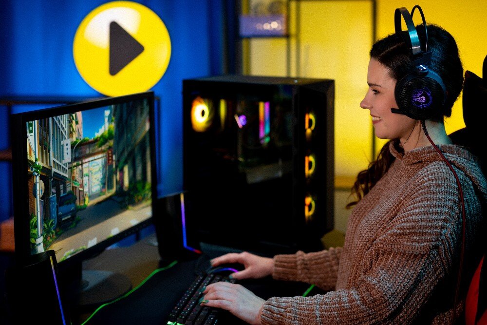 Komputer MAD DOG GeForce RTX5070 DLSS 4 Reflex Edition 2 STELLAR-A01DB32 Młoda kobieta w słuchawkach siedzi przy biurku i uśmiecha się podczas gry na komputerze, obok którego stoi przeszklona obudowa z widocznym kolorowym podświetleniem wentylatorów