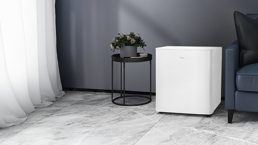 LODÓWKA TCL RF045DWE0  Na zdjęciu widać białą chłodziarkę TCL ustawiony obok granatowej sofy, na szarych płytkach imitujących marmur, przy ścianie w odcieniu grafitowym. Po lewej stronie stoi okrągły czarny stolik z donicą pełną kwiatów i filiżanką, a z boku opadają białe zasłony kompaktowa lodówka małe kuchnie biuro domek letniskowy elegancki design funkcjonalność wygoda