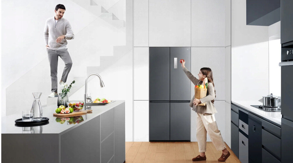 LODÓWKA TCL RC456CXE0 W przestronnej kuchni widać po lewej duży, szary półwysep z błyszczącym blatem, na którym stoją karafka z wodą, szklanki, deska z owocami i wazon z zielonymi gałązkami, a w tle mężczyzna schodzi po białych schodach. Po prawej stronie kadru kobieta z papierową torbą pełną warzyw i owoców unosi rękę w stronę lodówki French Door zabudowanej w wysokiej, jasnoszarej ścianie szafek inteligentne technologie cicha praca energooszczędność