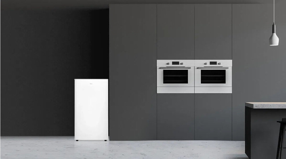 LODÓWKA TCL RF092DWE0 Na zdjęciu 2.jpg przedstawiono białą lodówkę wolnostojącą ustawioną przy ciemnej ścianie w minimalistycznej kuchni. Po prawej stronie widać wysoką zabudowę z dwoma piekarnikami w słupku oraz fragment wyspy kuchennej z betonowym blatem i zwisającą lampą mała lodówka kompaktowe wymiary do małej kuchni biuro domek letniskowy elegancki design funkcjonalne przechowywanie łatwa higiena wygoda użytkowania porządek komfort