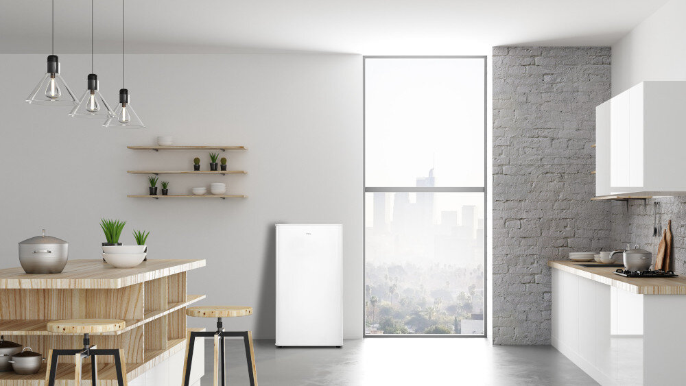 LODÓWKA TCL RF092DWE0 Na zdjęciu pokazano jasną, przestronną kuchnię z dużym oknem sięgającym od podłogi do sufitu, przez które widać panoramę miasta. Pośrodku przy ścianie stoi biała lodówka, a po bokach znajdują się wyspa kuchenna z drewnianym blatem oraz zabudowa z szafkami i płytą grzewczą kompaktowa lodówka funkcjonalność wygoda powłoka HIPS higiena regulowane nóżki stabilność półka XL dodatkowa przestrzeń regulowane półki miejsce na napoje małe kuchnie biuro domek letniskowy