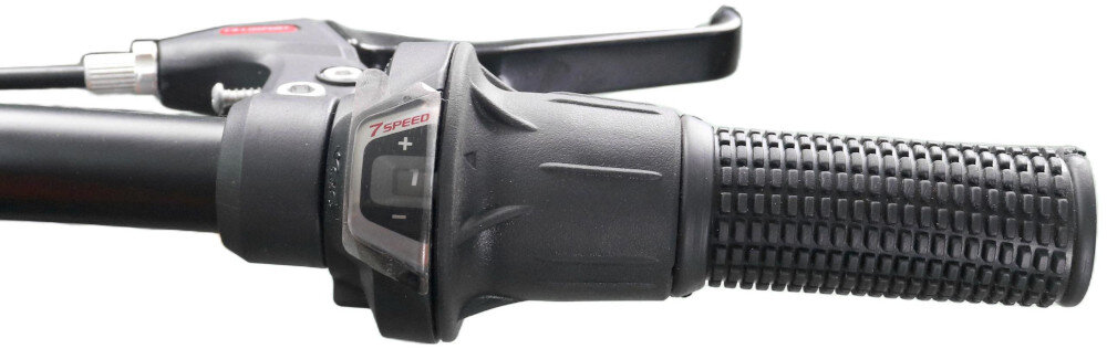Zbliżenie na czarną manetkę rowerową z widocznym napisem \'7 SPEED\' oraz oznaczeniami \'+\' i \'-\'.
