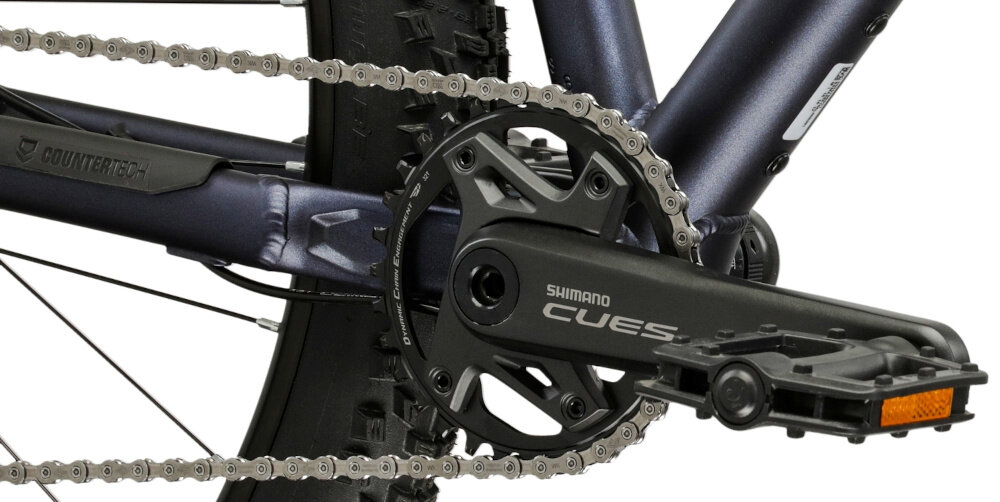 Zbliżenie na mechanizm korbowy roweru z napisem \'SHIMANO CUES\' na czarnej korbie. Widoczny jest również łańcuch oraz część ramy roweru w kolorze granatowo-grafitowym.