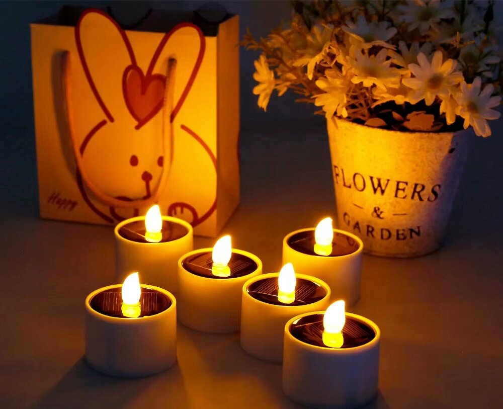 Na obrazie znajduje się sześć świec LED z efektem płomienia, ustawionych obok torby z rysunkiem królika i napisem \'Happy\'. W tle widoczna jest doniczka z napisem \'FLOWERS & GARDEN\' oraz białymi kwiatami.