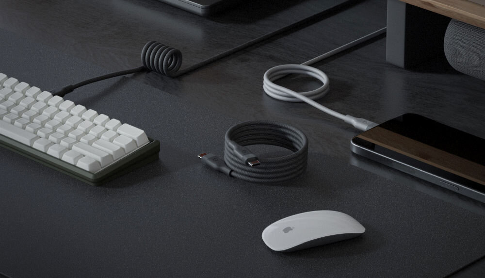 Na biurku znajduje się zwinięty czarny kabel USB-C - USB-C, klawiatura, mysz z logo w kształcie jabłka oraz smartfon. W tle widoczny jest laptop i inny kabel podłączony do urządzenia.