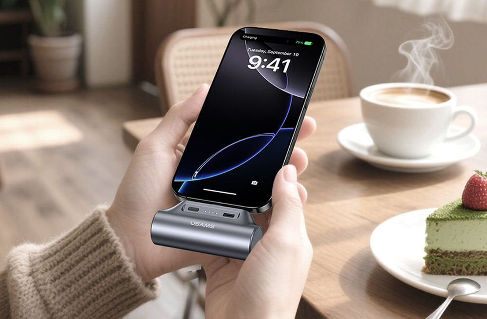 Osoba trzyma smartfon podłączony do powerbanku marki USAMS, na ekranie widoczna jest godzina 9:41 i data Tuesday, September 10. W tle znajduje się filiżanka kawy i kawałek ciasta na talerzu.