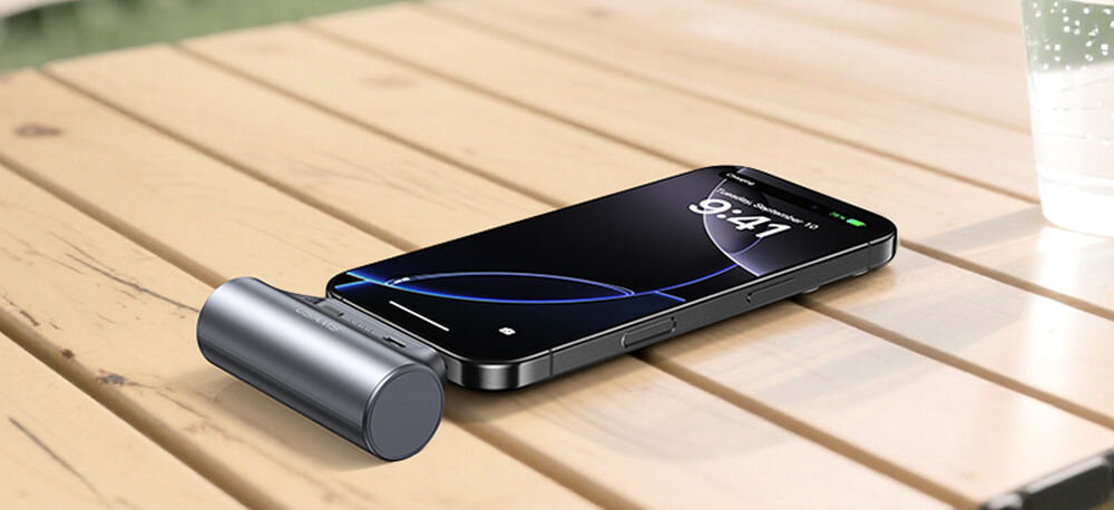 Na drewnianym stole leży smartfon z wyświetlaczem pokazującym godzinę 9:41, obok którego znajduje się srebrny powerbank o cylindrycznym kształcie. W tle widoczna jest szklanka z napojem.