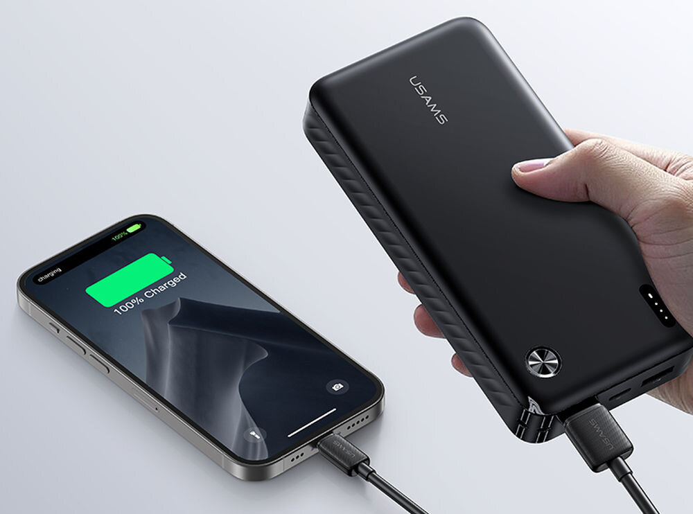 Na obrazie widać czarny powerbank z logo \'USAMS\' trzymany w dłoni, podłączony do smartfona, który pokazuje stan naładowania 100%.