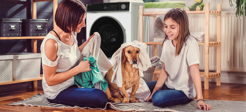 PRALKA SHARP ES-NFL714AWNNA-PL Na zdjęciu widać kobietę, dziewczynkę i psa siedzących na dywanie w domowym wnętrzu, bawiących się świeżo wypranym praniem. W tle, między nimi, ustawiona jest pralka SHARP, podkreślająca rodzinny i przytulny charakter sceny para higiena alergeny allergy smart 99,9% czystości