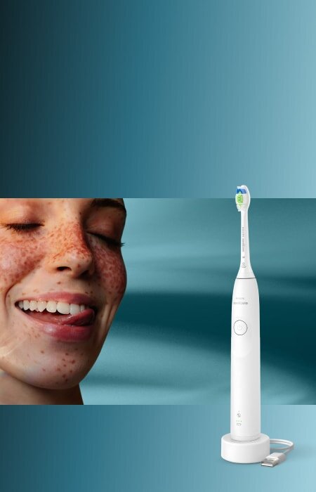 Zbliżenie na twarz uśmiechniętej kobiety, która językiem przesuwa po zębach. Obok szczoteczka soniczna.