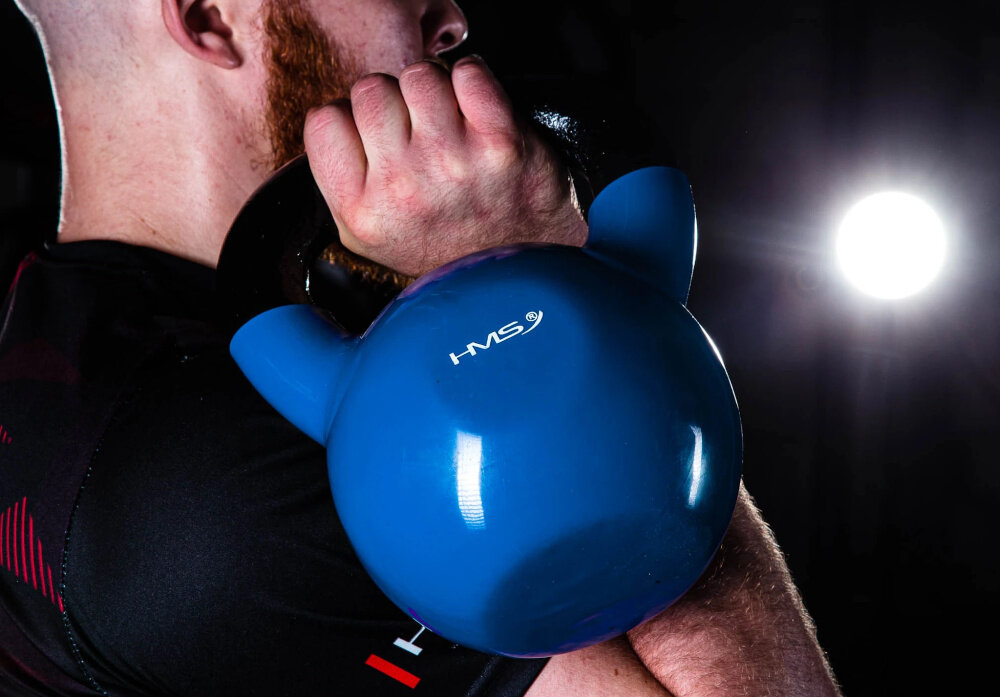 Kettlebell HMS KNV20 (20 kg) Niebiesko-czarny ciemne tło osoba trzyma kettlebell na ramieniu z wysokiej jakości żeliwa pokrytego warstwą niebieskiego winylu wytrzymały uchwyt malowany na czarno pewny chwyt minimalizuje ryzyko wyślizgnięcia się z dłoni