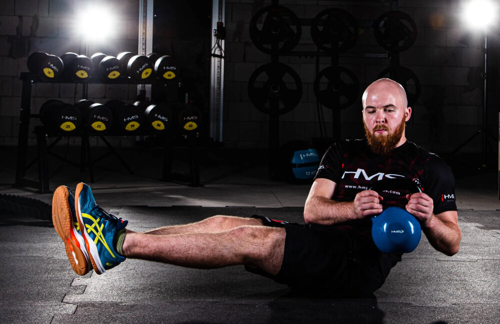 Kettlebell HMS KNV20 (20 kg) Niebiesko-czarny siłownia osoba ćwiczy skręty z kettlebellem siedząc na podłodze w tle sprzęty do ćwiczeń aktywują głębokie partie mięśniowe wsparcie w budowaniu siły funkcjonalnej oraz stabilności całego ciała zwiększa wydolność mięśni, poprawia postawę i ułatwia wykonywanie codziennych czynności