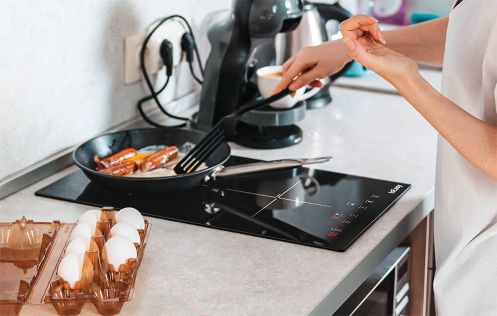 Płyta indukcyjna ISEASY LI2V-22 Osoba smaży jedzenie na patelni ustawionej na płycie indukcyjnej, obok leżą jajka w opakowaniu. W tle widać ekspres do kawy i inne kuchenne akcesoria blokada rodzicielska automatyczne wyłączanie bezpieczeństwo
