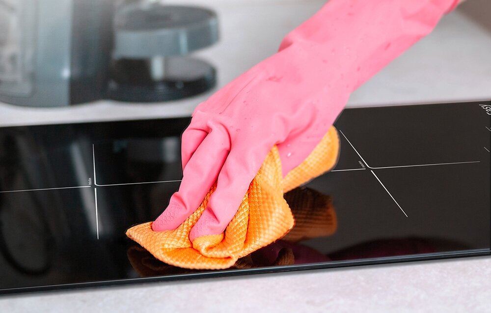 Płyta indukcyjna ISEASY LI2V-22 Dłoń w różowej rękawiczce przeciera płytę indukcyjną pomarańczową ściereczką. Zdjęcie podkreśla łatwość w utrzymaniu powierzchni w czystości cicha praca system chłodzenia łatwe czyszczenie