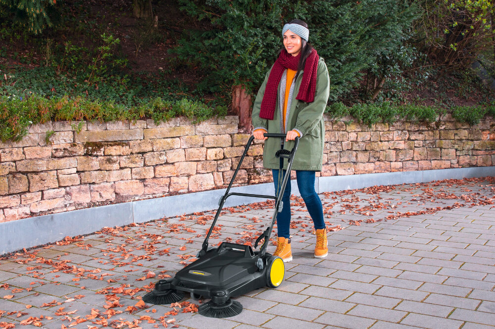 Zamiatarka KARCHER S 4 Twin Go Further 1.766-363.0 podjazd osoba zamiata podjazd za pomocą zamiatarki w tle murek z cegły drzewa krzewy rośliny zintegrowany zbiornik na zanieczyszczenia o pojemności 20 litrów możliwość łatwego wyjęcia i opróżnienia pojemnika bez kontaktu z brudem ogranicza unoszenie kurzu i rozsypywanie zanieczyszczeń