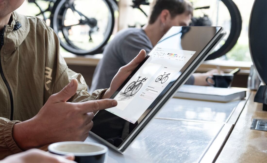 Laptop HP OmniBook w srebrnej obudowie na drewnianym stole w kawiarni, na ekranie kolorowa aplikacja do edycji graficznej z projektami i paletą kolorów, w tle osoba w czerwonej kurtce, obok kubek kawy i roślina doniczkowa, atmosfera kreatywnej pracy zdalnej.  