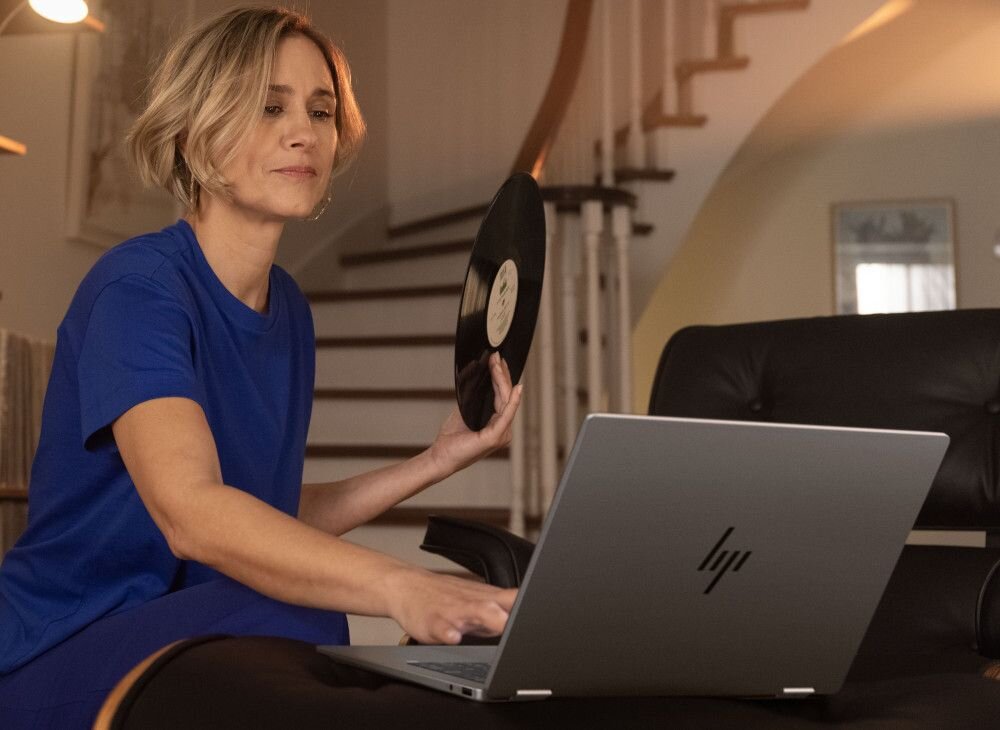 Kobieta w niebieskiej koszulce korzystająca z laptopa HP OmniBook i trzymająca płytę winylową, w nowoczesnym wnętrzu domowym, symbolizujący połączenie pracy, rozrywki i eleganckiego designu. - Certyfikat ENERGY STAR, EPEAT Gold z Climate+ 