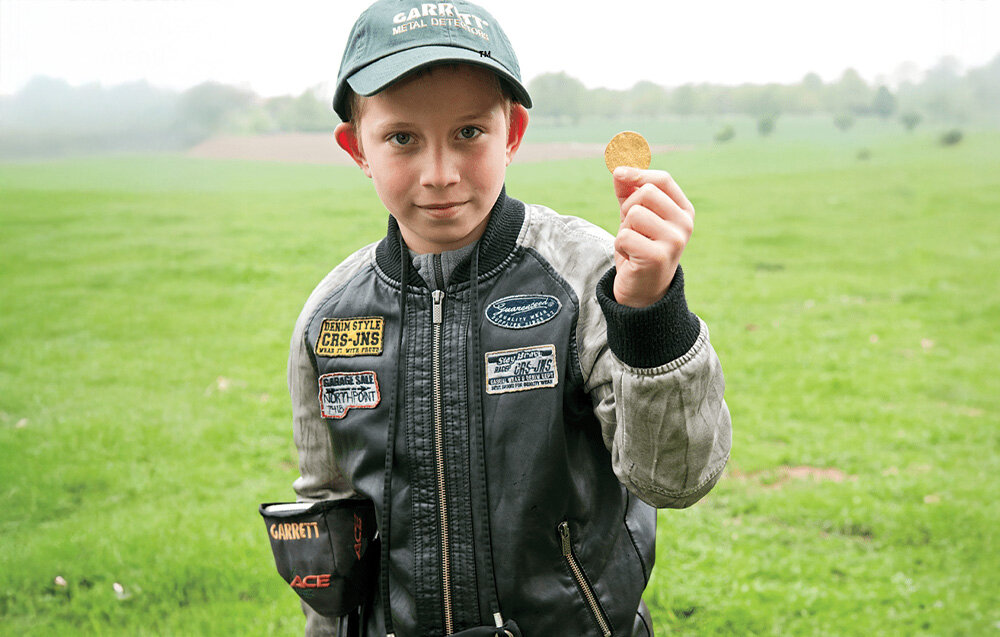 Wykrywacz metalu GARRETT ACE 250, głębokość, baterie, monety, kontrola, Chłopiec trzyma znalezioną monetę, ubrany w czapkę Garrett.