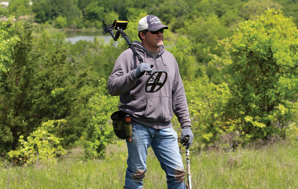 Wykrywacz metalu GARRETT ACE Apex, akumulator, litowo-jonowy, Micro-USB, czułość, Mężczyzna niesie wykrywacz metali na ramieniu w plenerze.