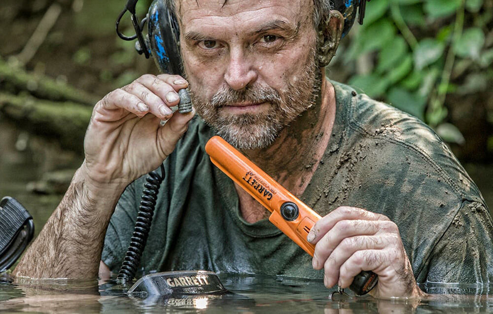 Osoba trzyma wykrywacz metalu GARRETT Pro-Pointer AT w wodzie. Widoczne jest logo \'GARRETT\' na urządzeniu oraz na słuchawkach.