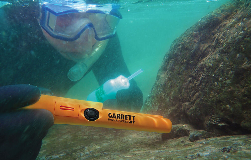 Osoba w masce do nurkowania trzyma wykrywacz metalu GARRETT Pro-Pointer AT pod wodą, obok skały. Na urządzeniu widoczne jest logo \'GARRETT Pro-Pointer AT\'.