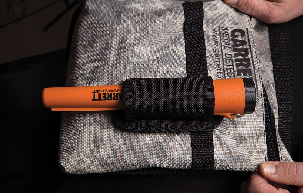 Pomarańczowy wykrywacz metalu z napisem \'GARRETT Pro-Pointer AT\' leży na szarej torbie z kamuflażowym wzorem i napisem \'GARRETT METAL DETECTORS www.garrett.com\'.