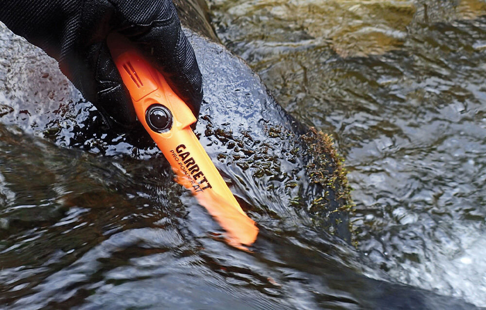 Osoba w rękawicy trzyma pomarańczowy wykrywacz metalu zanurzony w wodzie. Na urządzeniu widoczny jest napis \'GARRETT Pro-Pointer AT\'.