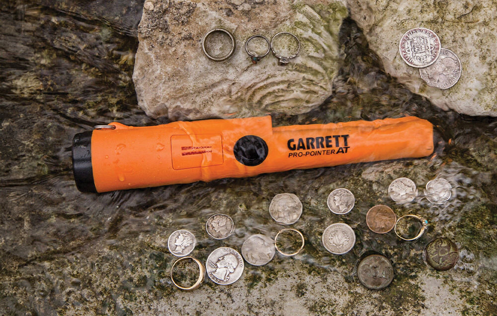Pomarańczowy wykrywacz metalu z napisem \'GARRETT Pro-Pointer AT\' leży na mokrych kamieniach, otoczony monetami i pierścionkami.