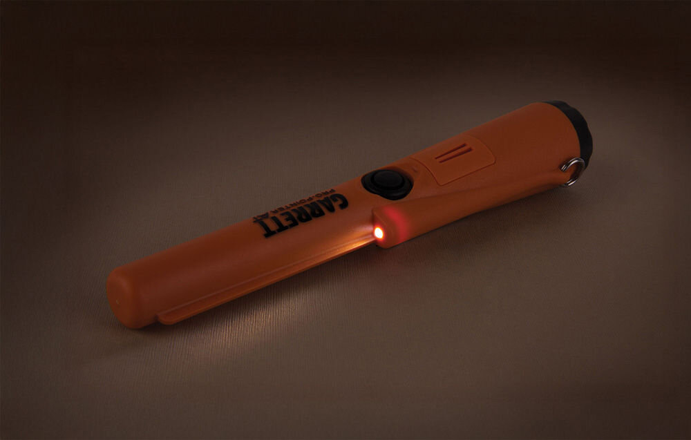 Pomarańczowy wykrywacz metalu z napisem \'GARRETT\' i świecącą czerwoną diodą. Urządzenie ma czarny przycisk i metalowy uchwyt na końcu.