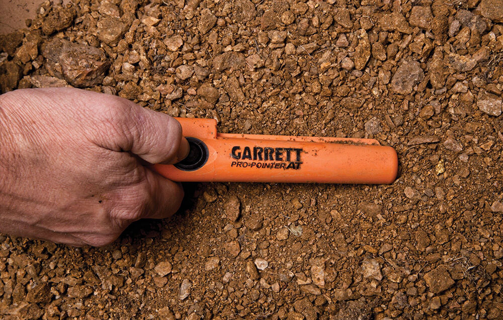 Pomarańczowy wykrywacz metalu z napisem \'GARRETT PRO-POINTER AT\' trzymany w dłoni na tle ziemi i kamieni.