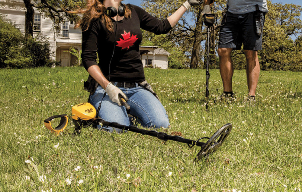 Wykrywacz metalu GARRETT ACE 300i, 5 trybów, Zero, Pinpointing, filtracja, Kobieta klęczy na łące z wykrywaczem Garrett ACE 300i obok niej.