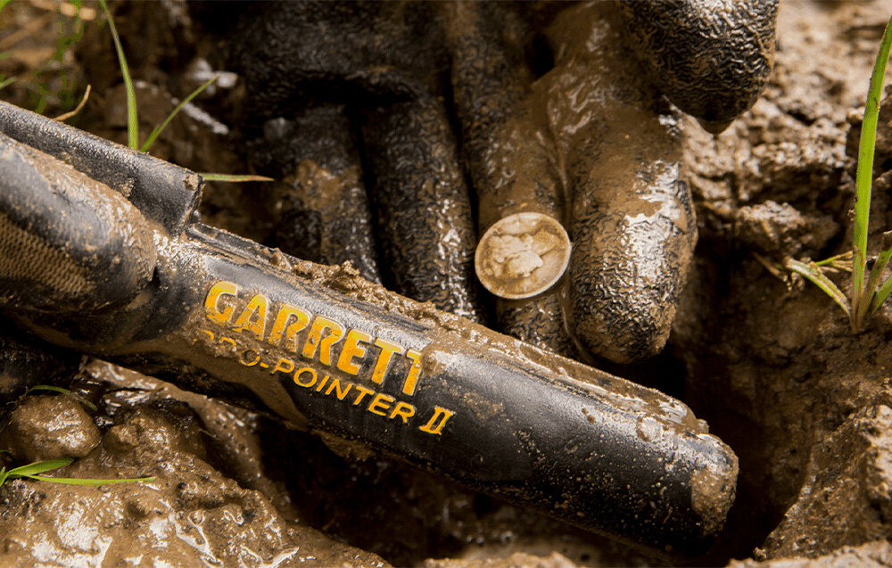 Wykrywacz metalu GARRETT Pro-Pointer II, odkrywanie, detekcja, skarby, sygnał, Dłoń w błocie ze srebrną monetą obok pinpointera Garrett.