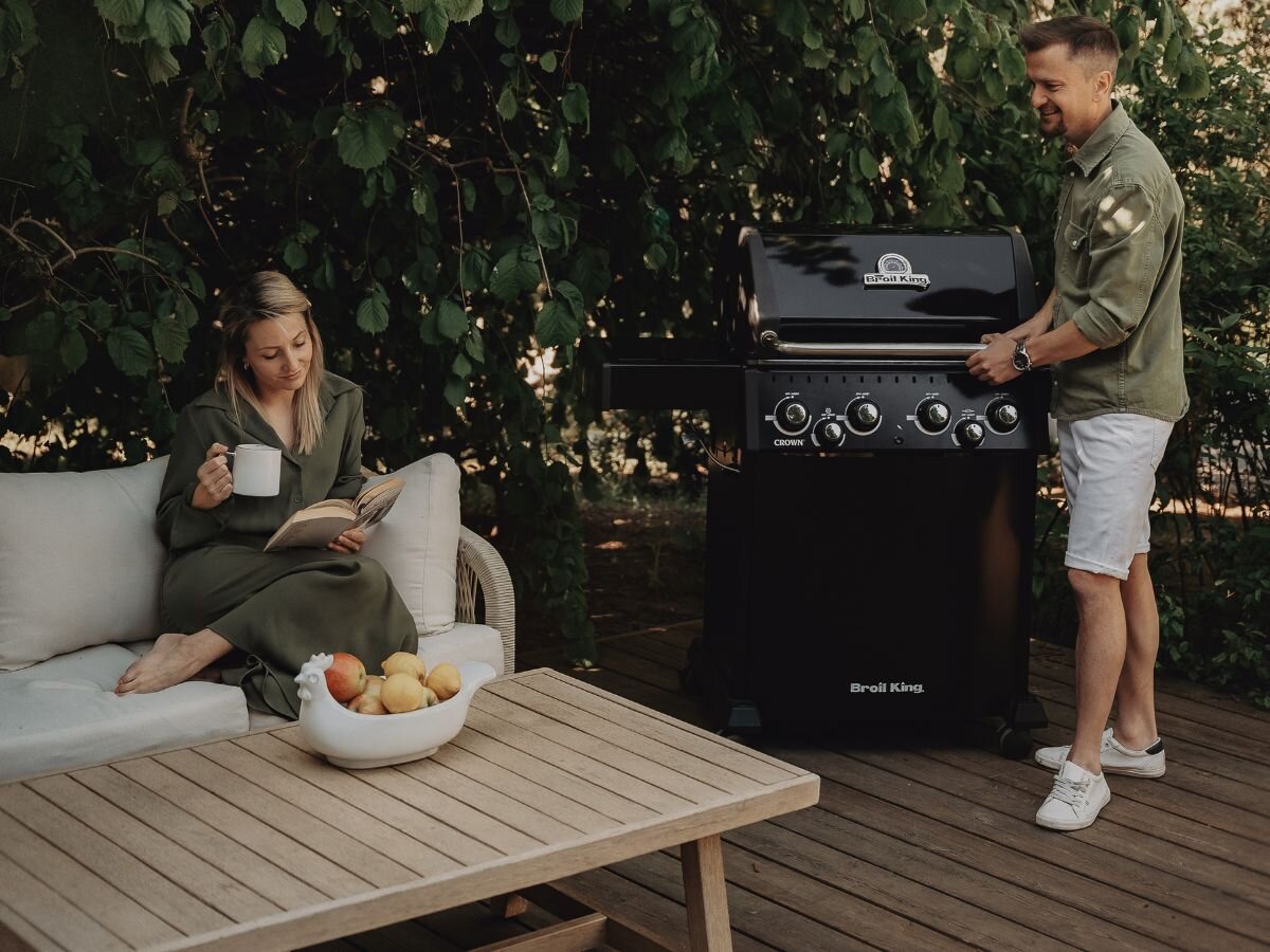 Osoba siedzi na sofie z książką i kubkiem, obok stoi grill gazowy Broil King z widocznym logo. Na stole przed sofą znajduje się misa z owocami.