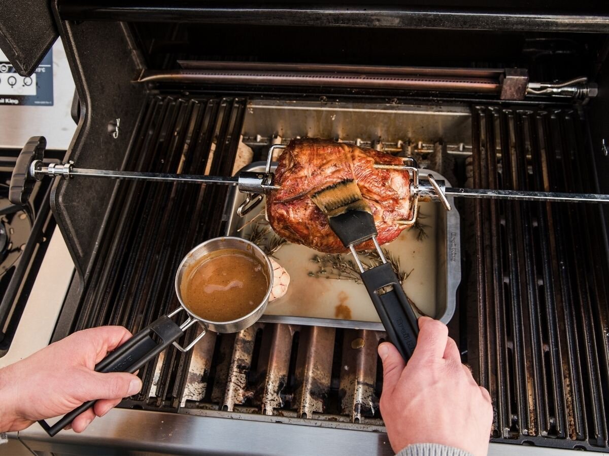 Osoba smaruje pieczone mięso na rożnie za pomocą pędzla, trzymając naczynie z sosem. Pod mięsem znajduje się brytfanna do grillowania z logo Broil King.