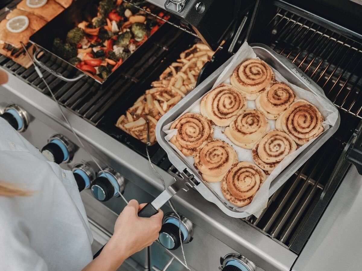 Osoba trzyma brytfannę z bułeczkami cynamonowymi na grillu, obok znajdują się grillowane warzywa i frytki. Widoczne są pokrętła do regulacji temperatury na grillu.