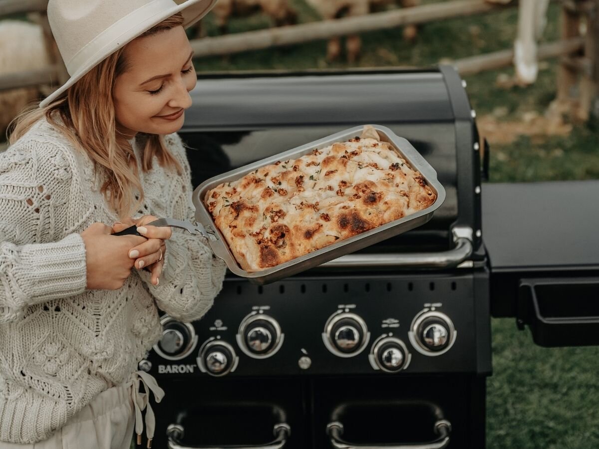Osoba trzyma brytfannę z zapiekanym daniem przed grillem z widocznymi pokrętłami. Na grillu widoczne jest logo \'BARON\'.