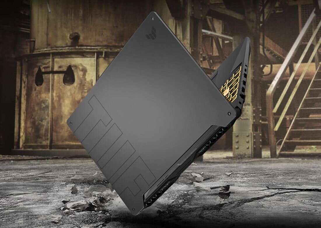 Zamknięty laptop ASUS TUF Gaming A15 w kolorze grafitowym, ustawiony pod kątem na pękniętej betonowej powierzchni w industrialnym otoczeniu, widoczne tłoczenie „TUF” na pokrywie i charakterystyczny wzór, fragment podświetlanej klawiatury. - MIL-STD-810H 