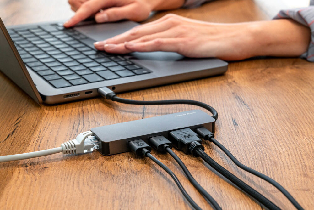 Hub USB XQUANTUM UH220 5w1 na biurku z podłączonymi przewodami w tle osoba pisząca na komputerze