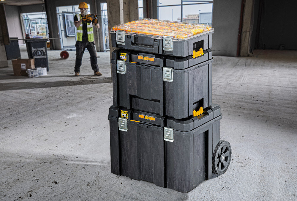 Skrzynka narzędziowa DEWALT DWST83346-1 magazyn plac budowy skrzynka ustawiona na innych skrzynkach w tle pracuje osoba otwór umożliwiający założenie kłódki bezpieczeństwo przechowywanych narzędzi ochrona przed dostępem osób trzecich