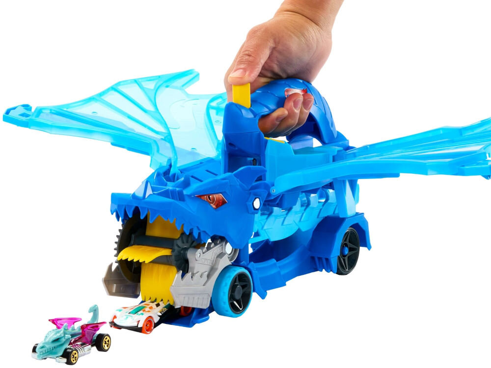 Zestaw pojazdów Hot Wheels City Transporter podwójny smok JBM72 Ręka trzyma niebieski pojazd Hot Wheels w kształcie smoka ze skrzydłami, który ma otwartą paszczę, z której wyjeżdżają dwa małe samochodziki.