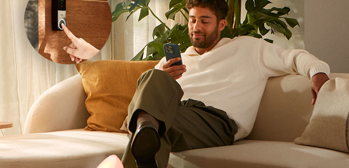 Wideodzwonek PHILIPS HUE 929003816503 Bezprzewodowy Wi-Fi Biało-czarny, powiadomienie, podgląd, na żywo, gość, Mężczyzna na kanapie korzystający ze smartfona, w kółku powiększone zdjęcie palca naciskającego przycisk wideodomofonu Hue Secure.