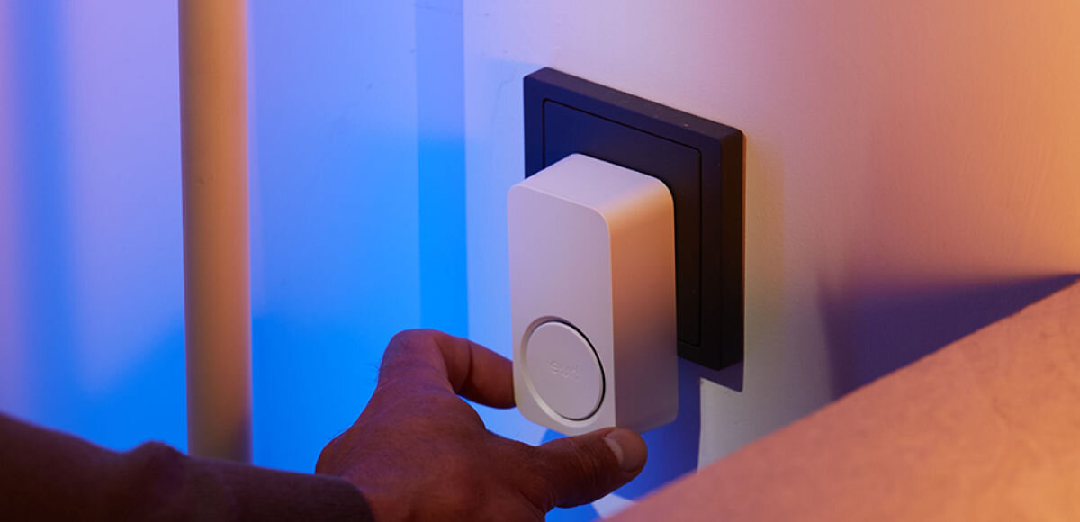 Dzwonek PHILIPS HUE, gniazdko, kable, skomplikowane, wiercenie, Zbliżenie na rękę wkładającą sygnalizator Hue Secure do gniazdka w ścianie, tonacja światła niebiesko-pomarańczowa.