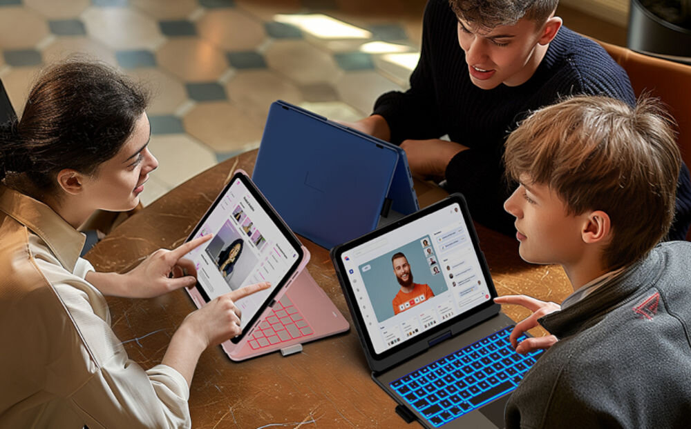 Trzy osoby siedzą przy stole, każda z nich używa tabletu z klawiaturą w etui. Na ekranach widoczne są różne aplikacje, w tym przeglądarka internetowa i komunikator.