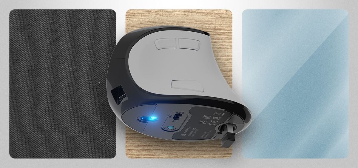 Na obrazie widoczna jest biało-czarna mysz komputerowa z kilkoma przyciskami na górze i niebieskim podświetleniem na spodzie. Mysz leży na drewnianej powierzchni, obok dwóch prostokątnych paneli – czarnego i niebieskiego.