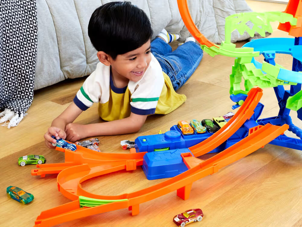 Tor Hot Wheels Stunt Tracks Powietrzna kraksa JFD12 Chłopiec w żółtej koszulce z uśmiechem leży na drewnianej podłodze i bawi się zestawem torów Hot Wheels. Na torze startowym leży kilka autek. Chłopiec trzyma w dłoni niebieski samochodzik i kieruje go na tor. W tle widoczne łóżko i narzuta.